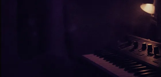 Arbeitsplatz bei Nacht: eine Lampe ausserhalb des Bildes, ein Synthesizer am Rand, der Rest im violetten Dunkel.